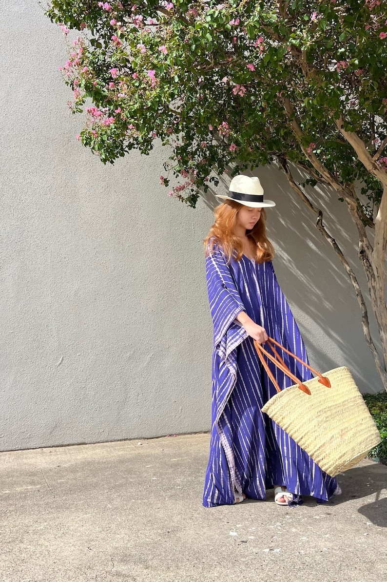 May include: A woman wearing a blue and white striped caftan, a white hat with a black band, and white sandals. She is holding a large straw tote bag with brown leather handles. The background includes a tree with pink flowers and a gray wall.