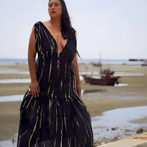 May include: A black maxi dress with a plunging neckline and a flowing skirt. The dress features vertical white streaks and is worn outdoors near the water. The model is standing on a concrete ledge.