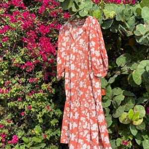 May include: A long, orange floral dress with white leaf print. The dress has a V-neckline and 3/4 sleeves. It is hanging against a backdrop of pink flowers.