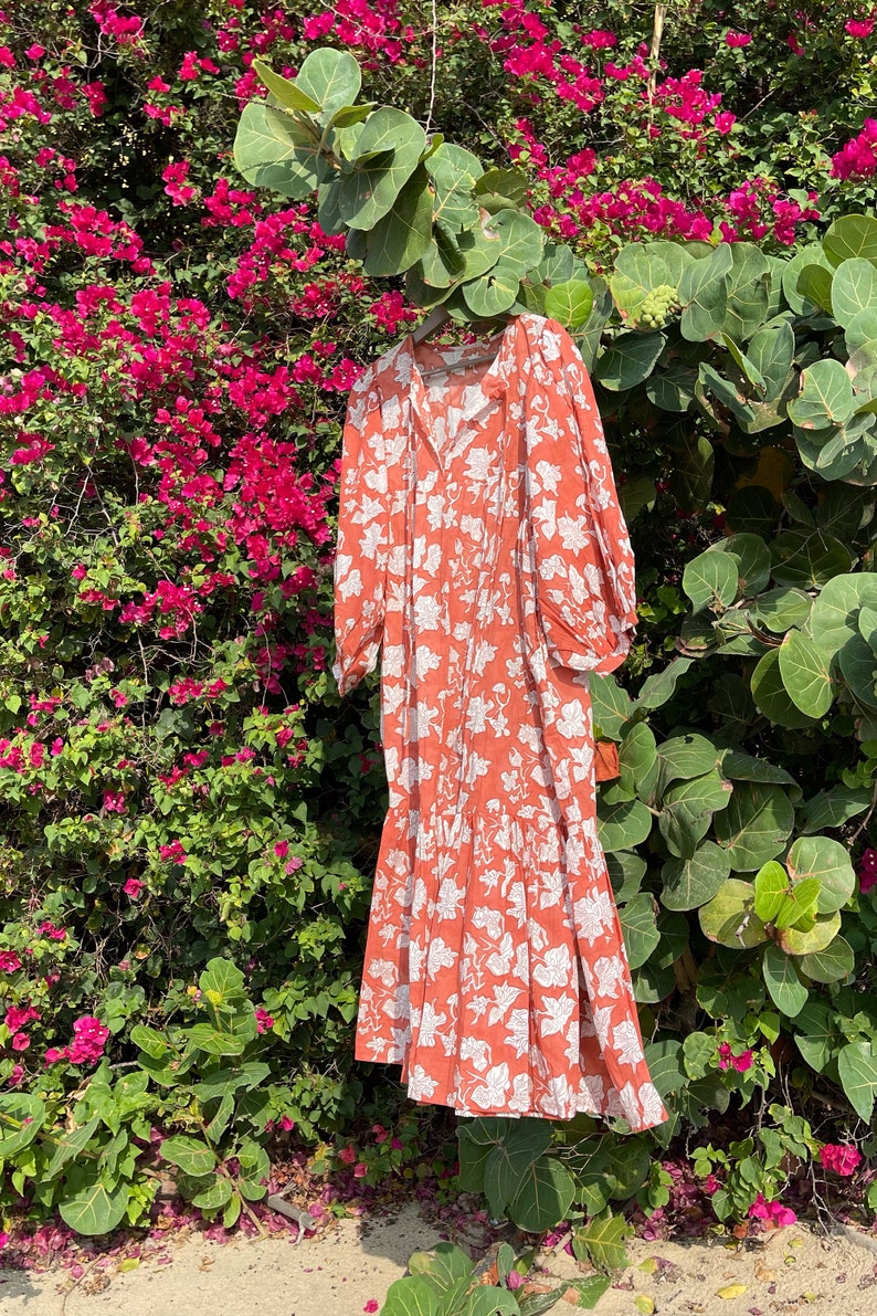 May include: A long, orange floral dress with white leaf print. The dress has a V-neckline and 3/4 sleeves. It is hanging against a backdrop of pink flowers.