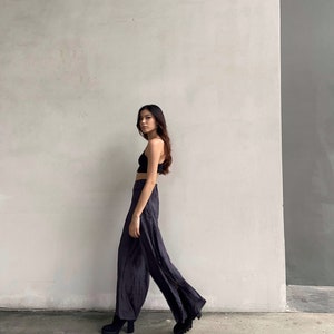 Puede incluir: Una mujer con un top negro corto y pantalones negros anchos con una abertura lateral. Est&aacute; de pie frente a una pared blanca con una secci&oacute;n gris a la derecha.