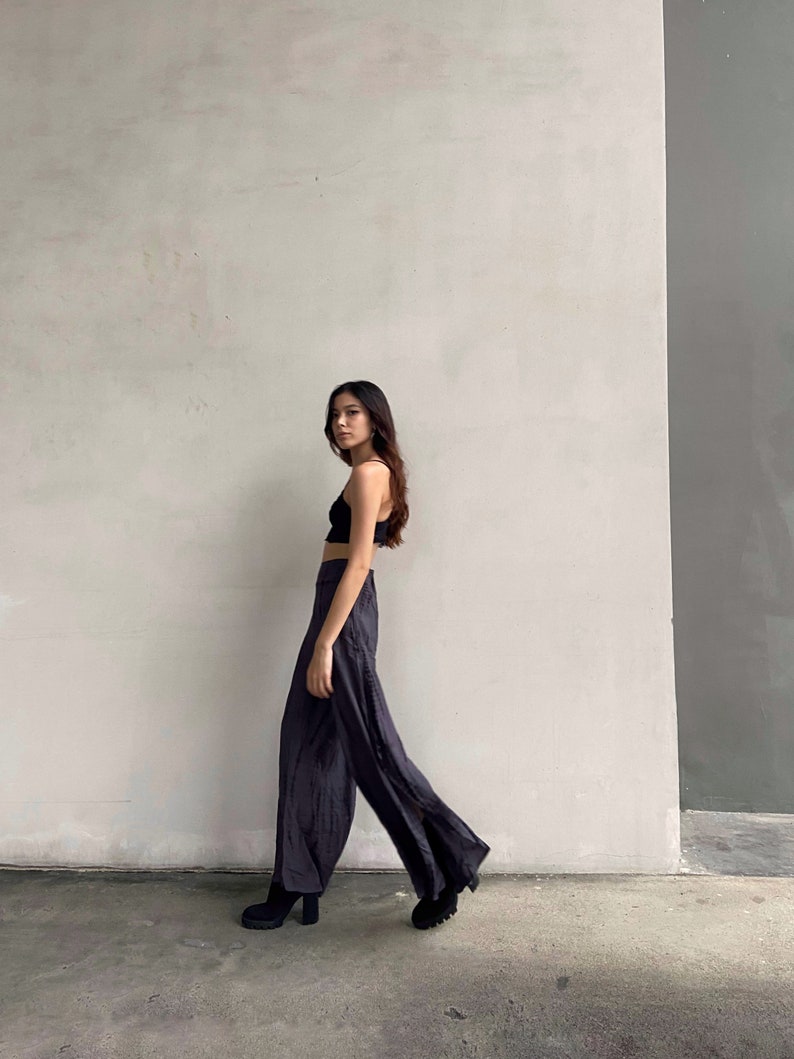 Puede incluir: Una mujer con un top negro corto y pantalones negros anchos con una abertura lateral. Est&aacute; de pie frente a una pared blanca con una secci&oacute;n gris a la derecha.