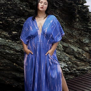 Op de afbeelding: Een vrouw draagt een lange, blauwe en roze tie-dye maxi-jurk met een diepe V-hals en zijsplitten. De jurk heeft een getailleerde taille en zakken.