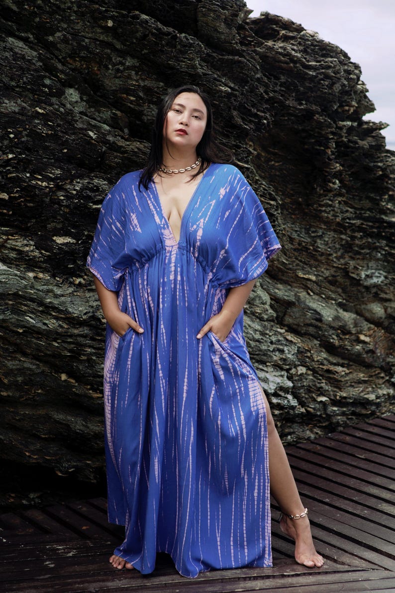 Op de afbeelding: Een vrouw draagt een lange, blauwe en roze tie-dye maxi-jurk met een diepe V-hals en zijsplitten. De jurk heeft een getailleerde taille en zakken.