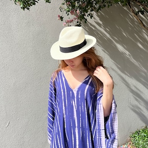 May include: A woman's summer outfit featuring a white straw fedora hat with a black band, and a blue and white striped caftan dress. The outfit is set against a gray wall with a tree and pink flowers.