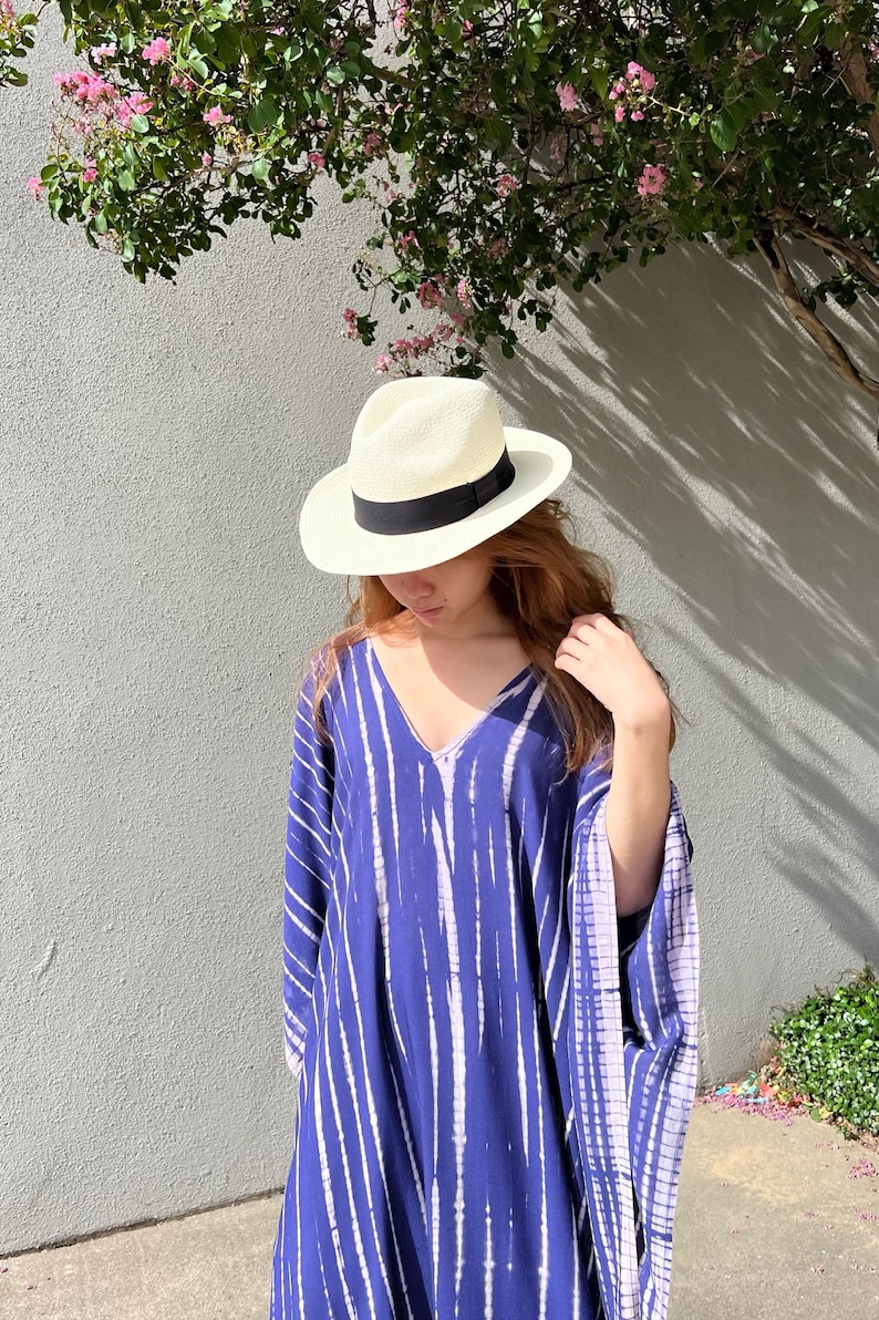 May include: A woman's summer outfit featuring a white straw fedora hat with a black band, and a blue and white striped caftan dress. The outfit is set against a gray wall with a tree and pink flowers.