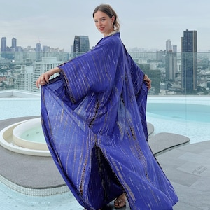 May include: A woman wearing a long, flowing purple and brown tie-dye kimono with a side slit. The kimono is made of a lightweight fabric and has a relaxed fit. The woman is standing on a rooftop with a city skyline in the background.