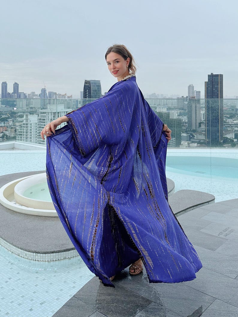 May include: A woman wearing a long, flowing purple and brown tie-dye kimono with a side slit. The kimono is made of a lightweight fabric and has a relaxed fit. The woman is standing on a rooftop with a city skyline in the background.