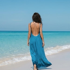 Woman wearing a blue turquoise open back maxi dress with tie shoulders and a loose flowy silhouette on a beach, styled as resort wear for vacation.