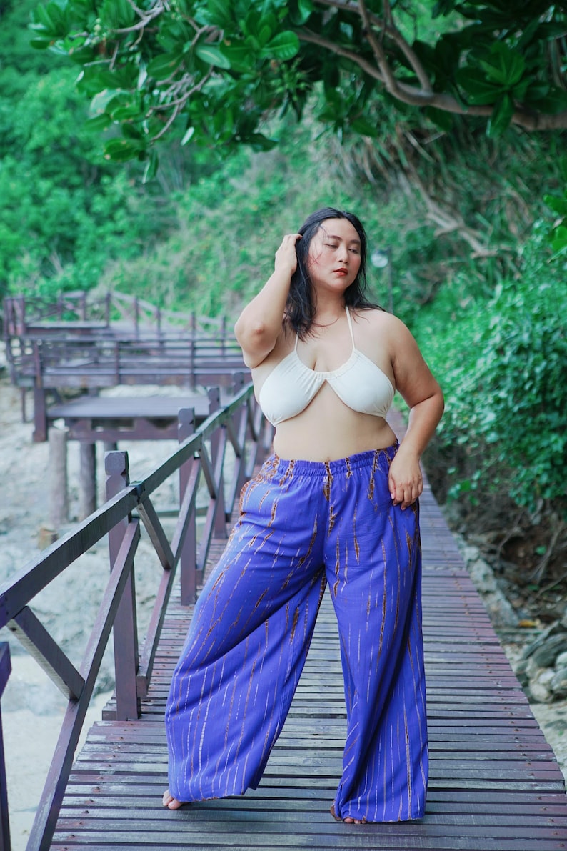 May include: A woman wearing a white halter top and purple wide-leg pants with a gold pattern stands on a wooden walkway.