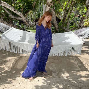 May include: A long, flowing navy blue dress with white vertical stripes. The dress has a V-neck and wide sleeves. A white macrame hammock hangs in the background, with trees and sand surrounding the scene.