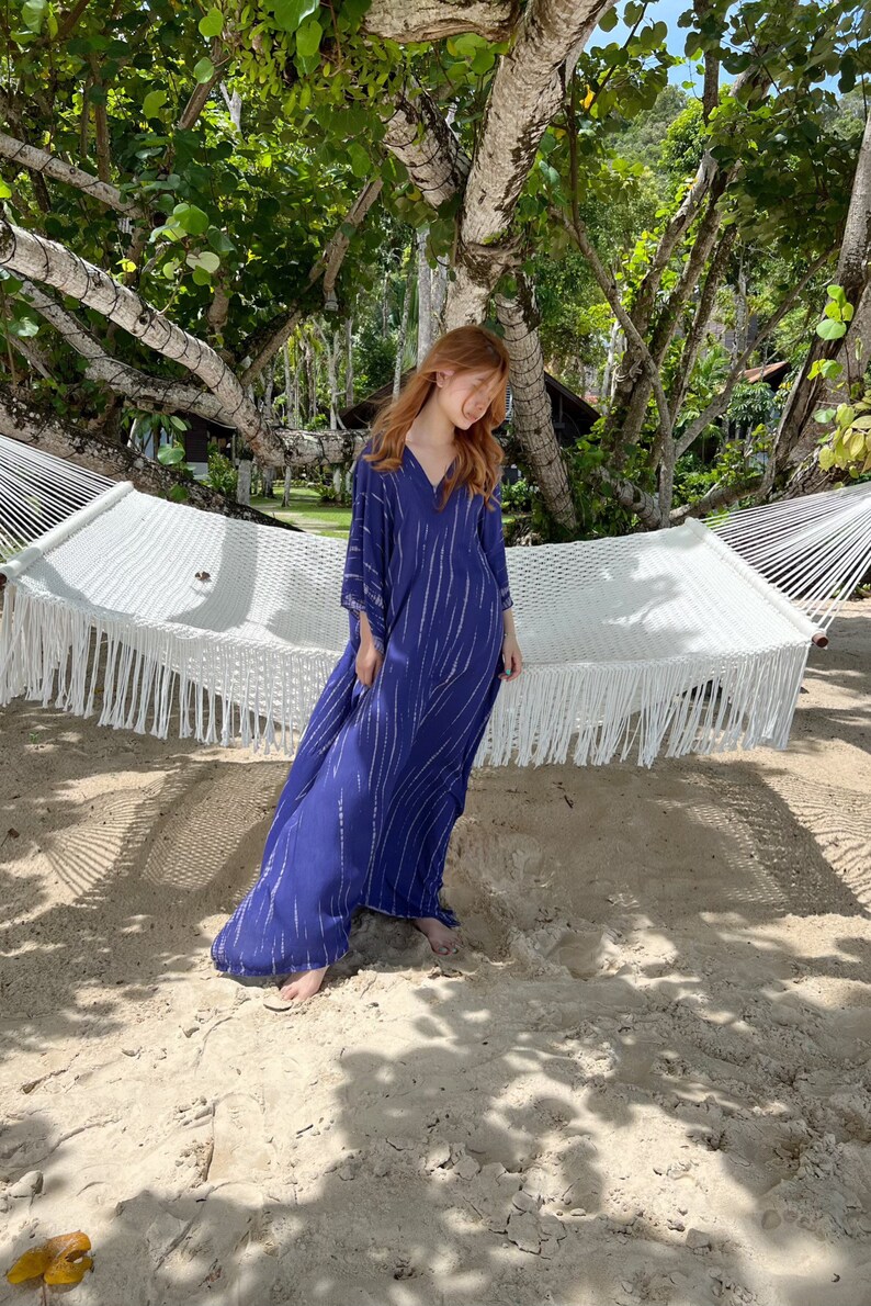 May include: A long, flowing navy blue dress with white vertical stripes. The dress has a V-neck and wide sleeves. A white macrame hammock hangs in the background, with trees and sand surrounding the scene.