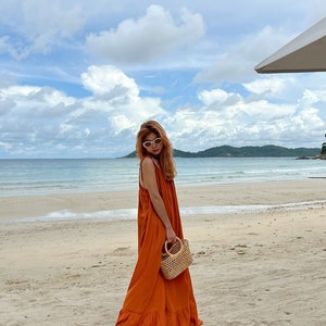 An orange maxi dress with a flowing silhouette. The dress is sleeveless and worn on a sandy beach. A small, woven handbag with a wooden handle is held. The background features a beach, ocean, and cloudy sky.
