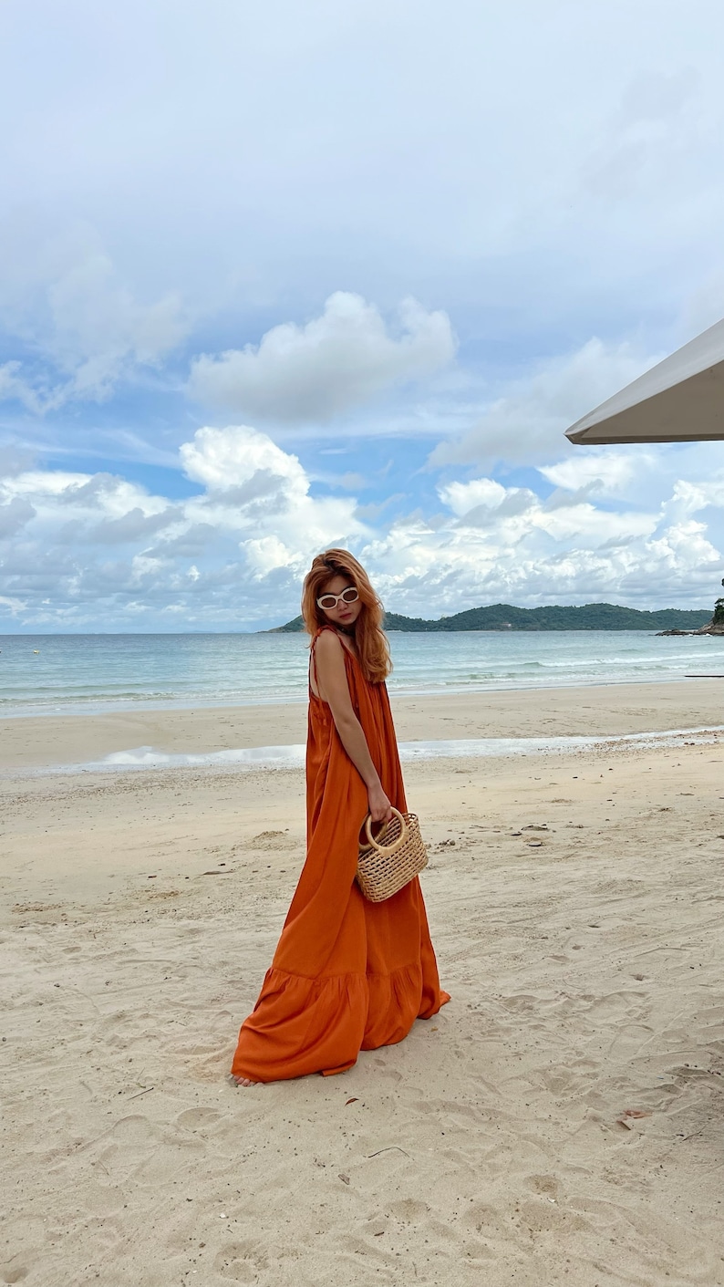 An orange maxi dress with a flowing silhouette. The dress is sleeveless and worn on a sandy beach. A small, woven handbag with a wooden handle is held. The background features a beach, ocean, and cloudy sky.