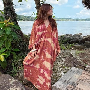 May include: A long, flowing dress with a V-neck and tie-dye pattern in shades of red, orange, and cream. The dress has long sleeves and a relaxed fit. The model is standing near the water, wearing the dress.