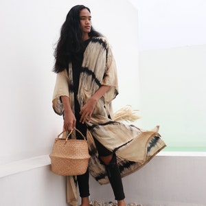 May include: A person wearing a long, beige and black tie-dye kimono with black pants and a woven straw tote bag.