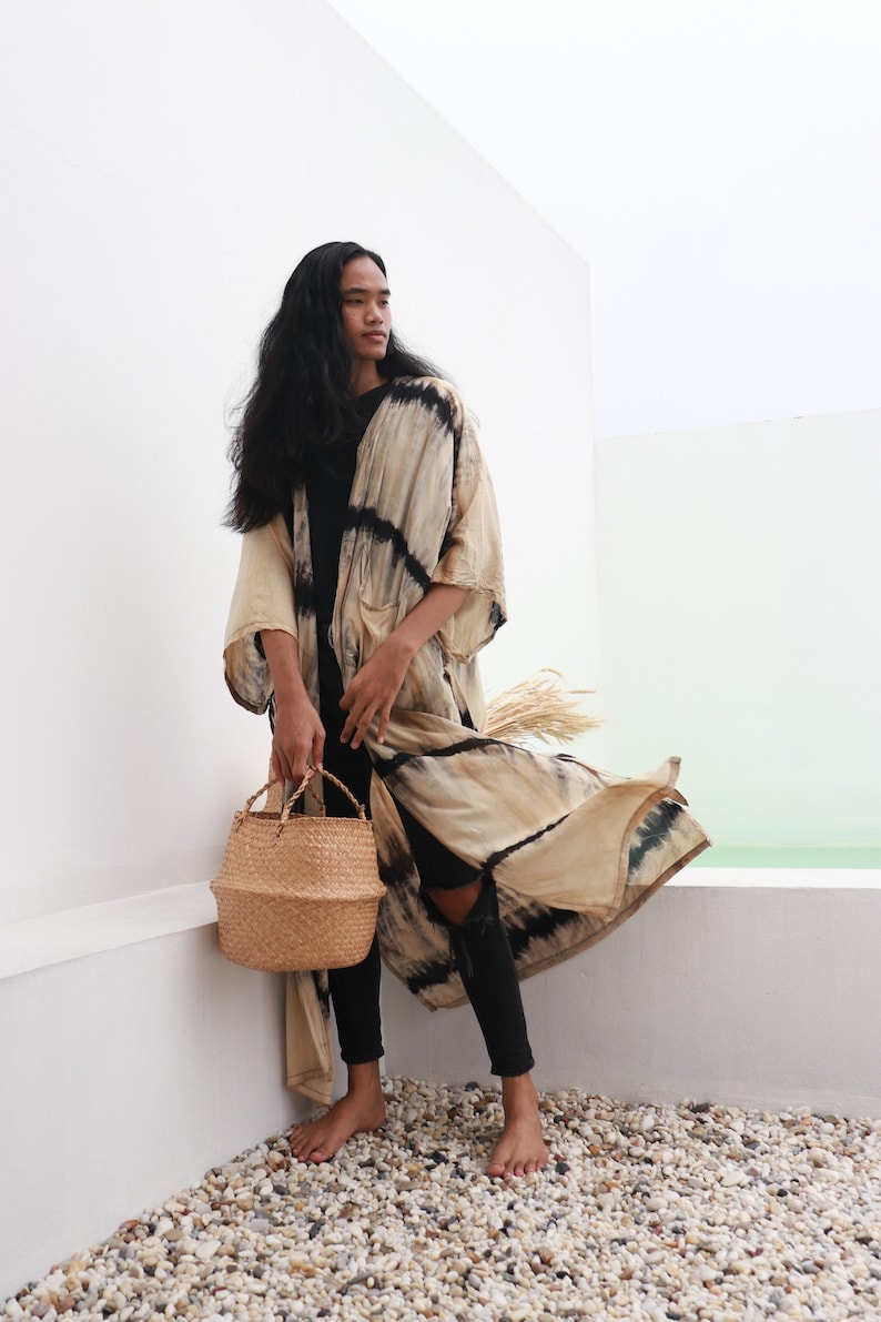 May include: A person wearing a long, beige and black tie-dye kimono with black pants and a woven straw tote bag.