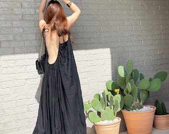 May include: A black maxi dress with a low back and thin straps. The dress is made of a soft, flowing fabric and has a tiered skirt. The model is wearing the dress with a gold bracelet and a black purse.