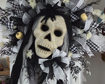 May include: A Halloween wreath featuring a skull with a black spider on its head. The wreath is decorated with black and white ribbons, faux flowers, and small pumpkins. The color scheme is black, white, and cream.