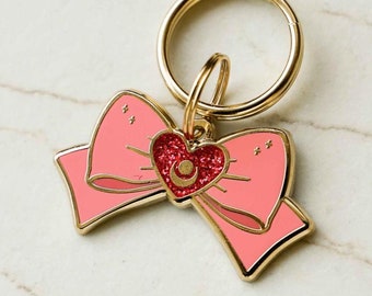 May include: A pink bow-shaped keychain with a gold-colored ring. The bow has a gold outline and a red heart in the center. The heart has a crescent moon design. The keychain is on a white marble surface.