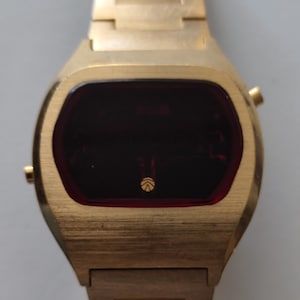May include: A gold-toned digital wristwatch featuring a brushed metal finish and a dark red rectangular display screen. The watch face includes a small circular logo at the bottom center. It has a matching gold-toned metal link bracelet strap.