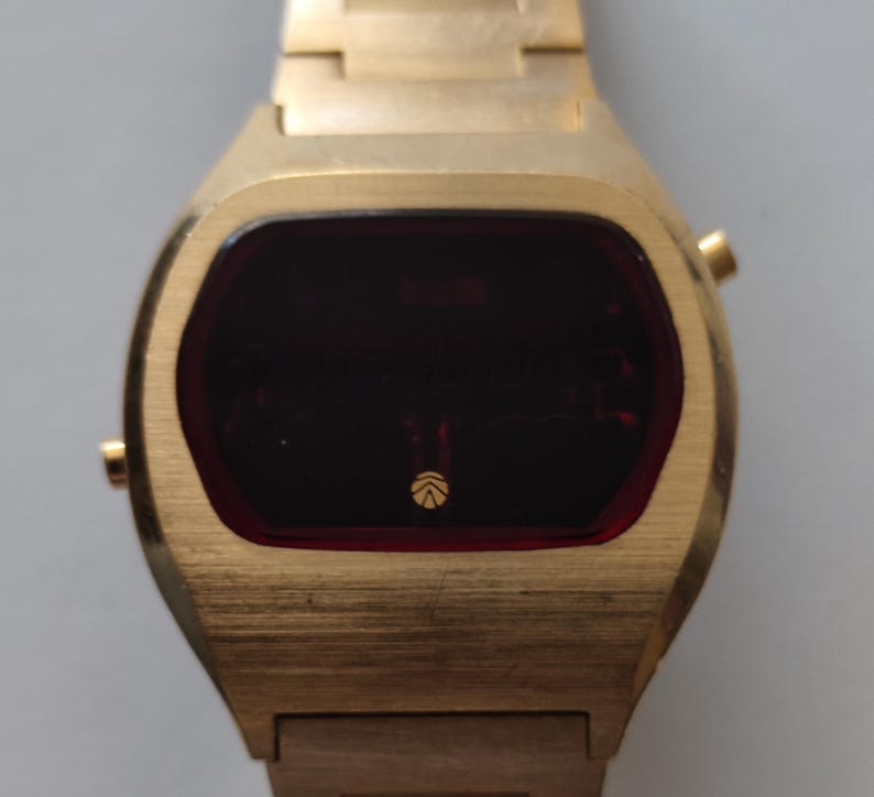 May include: A gold-toned digital wristwatch featuring a brushed metal finish and a dark red rectangular display screen. The watch face includes a small circular logo at the bottom center. It has a matching gold-toned metal link bracelet strap.