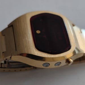 May include: A gold-tone digital wristwatch featuring a brushed metal finish, a dark red rectangular display screen with a small circular logo, and a matching link bracelet. Two silver-colored circular buttons are positioned on the side of the case.