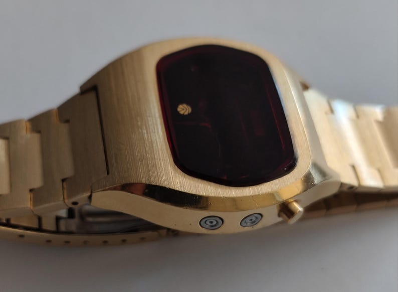 May include: A gold-tone digital wristwatch featuring a brushed metal finish, a dark red rectangular display screen with a small circular logo, and a matching link bracelet. Two silver-colored circular buttons are positioned on the side of the case.