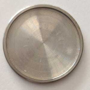 May include: A circular, metallic watch case back with a brushed silver finish. The component features a smooth, slightly domed center and a raised outer rim, designed to seal the movement of a timepiece. The surface reflects light in a radial pattern.