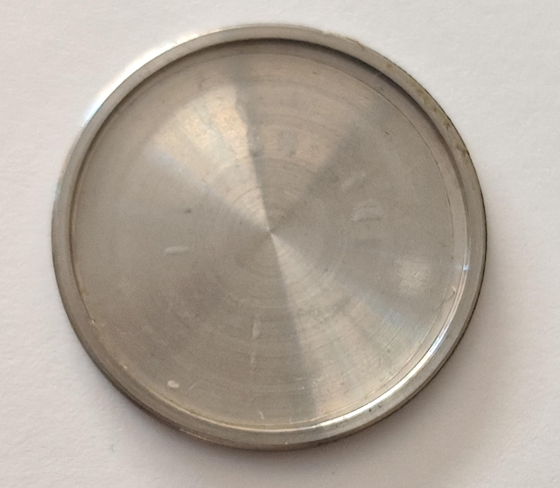 May include: A circular, metallic watch case back with a brushed silver finish. The component features a smooth, slightly domed center and a raised outer rim, designed to seal the movement of a timepiece. The surface reflects light in a radial pattern.