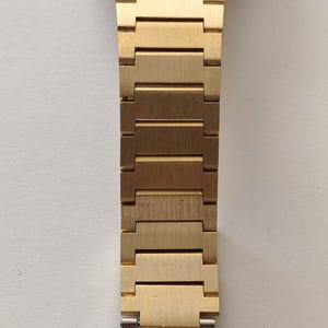 May include: A gold-toned metal watch band featuring a series of rectangular, brushed-finish links that connect to a polished silver-toned metal clasp at the bottom. The band has a uniform width and a structured, segmented design.