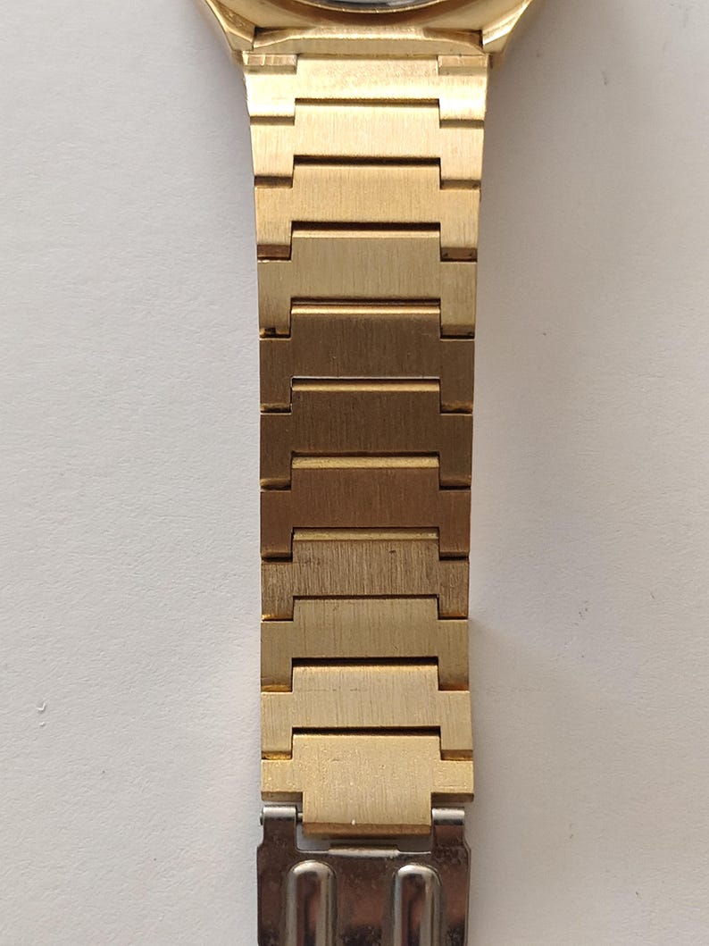 May include: A gold-toned metal watch band featuring a series of rectangular, brushed-finish links that connect to a polished silver-toned metal clasp at the bottom. The band has a uniform width and a structured, segmented design.