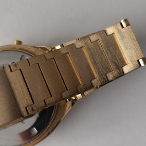 May include: A gold-toned metal watch bracelet with a brushed finish, featuring rectangular interlocking links. The bracelet is attached to a round watch case with a silver-colored circular back plate visible, showing a small gold-toned crown on the side.