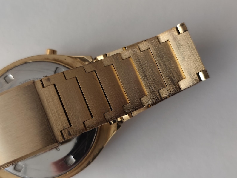 May include: A gold-toned metal watch bracelet with a brushed finish, featuring rectangular interlocking links. The bracelet is attached to a round watch case with a silver-colored circular back plate visible, showing a small gold-toned crown on the side.