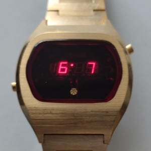 May include: A gold-tone digital wristwatch with a rectangular case and a matching metal link bracelet. The watch face features a dark red screen displaying the time in bright red LED numerals, with a small circular logo positioned below the display.