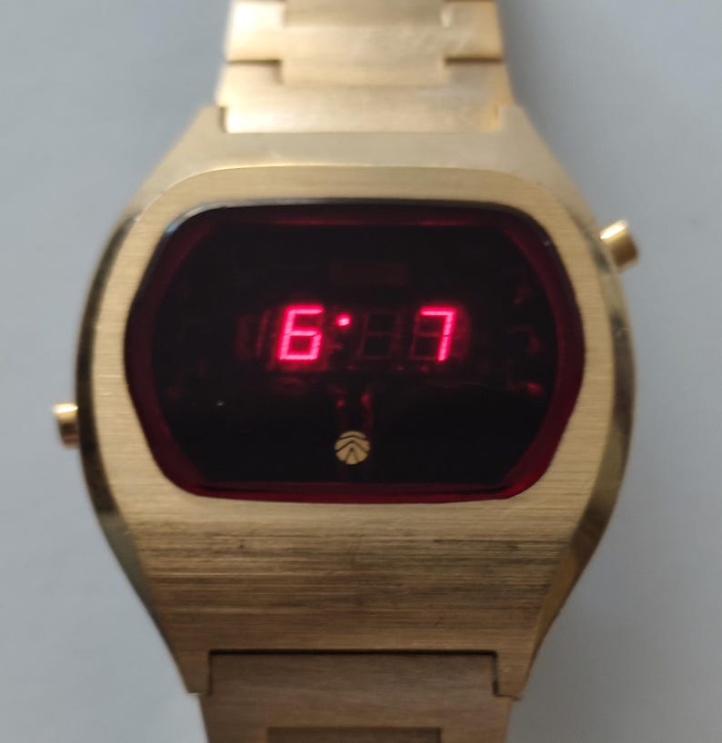 May include: A gold-tone digital wristwatch with a rectangular case and a matching metal link bracelet. The watch face features a dark red screen displaying the time in bright red LED numerals, with a small circular logo positioned below the display.