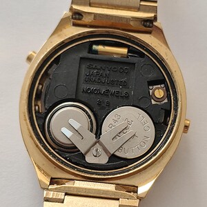 May include: A gold-tone wristwatch with the back casing removed, revealing the internal mechanism. The black plastic movement plate is embossed with SANYO CO JAPAN UNADJUSTED NO JEWELS. Two silver button cell batteries are secured by a metal clip.