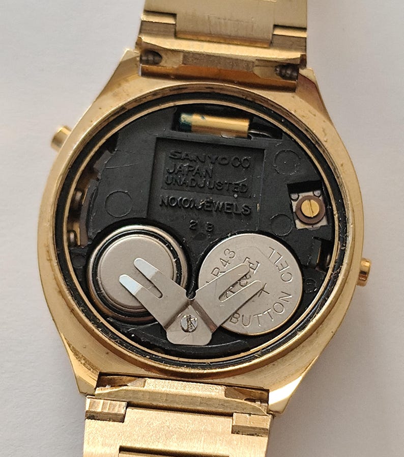 May include: A gold-tone wristwatch with the back casing removed, revealing the internal mechanism. The black plastic movement plate is embossed with SANYO CO JAPAN UNADJUSTED NO JEWELS. Two silver button cell batteries are secured by a metal clip.