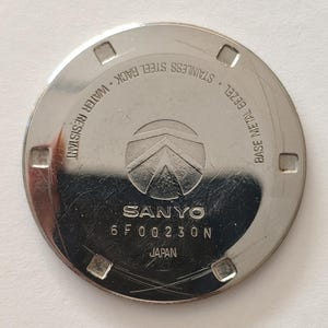 May include: A circular silver-toned stainless steel watch case back featuring the SANYO logo, the text 6 F 00 230 N, JAPAN, BASE METAL BEZEL, STAINLESS STEEL BACK, and WATER RESISTANT. The surface has four square notches along the outer edge.