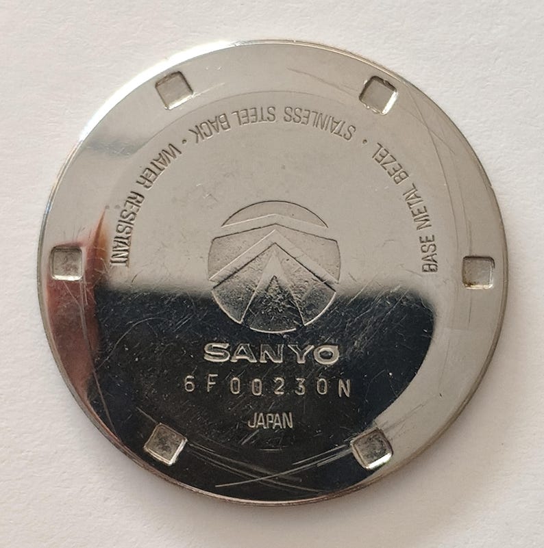 May include: A circular silver-toned stainless steel watch case back featuring the SANYO logo, the text 6 F 00 230 N, JAPAN, BASE METAL BEZEL, STAINLESS STEEL BACK, and WATER RESISTANT. The surface has four square notches along the outer edge.