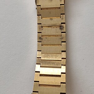 May include: A gold-tone metal watch band featuring a series of rectangular, interlocking links with a brushed finish. The band is attached to a watch case with a circular, silver-colored back. The metal shows signs of wear and minor surface discoloration.