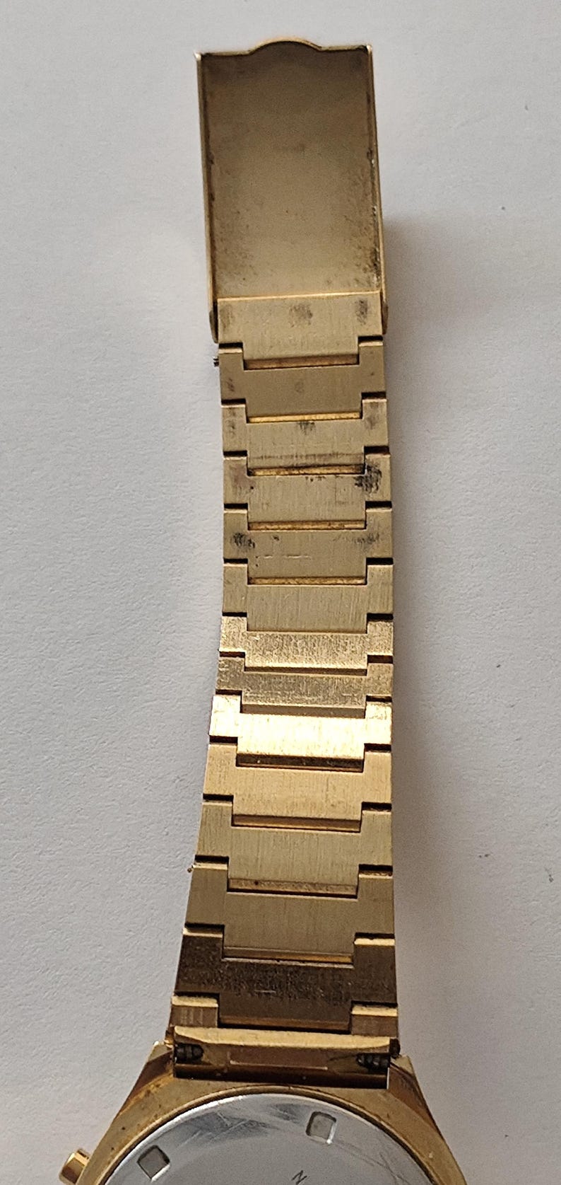 May include: A gold-tone metal watch band featuring a series of rectangular, interlocking links with a brushed finish. The band is attached to a watch case with a circular, silver-colored back. The metal shows signs of wear and minor surface discoloration.