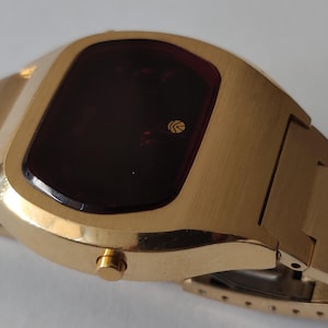 May include: A gold-tone digital wristwatch featuring a rectangular case with rounded edges and a dark red display screen. The watch includes a matching gold-tone metal link bracelet and a single circular side button for operation.
