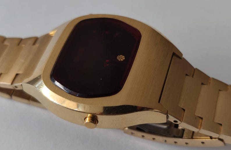 May include: A gold-tone digital wristwatch featuring a rectangular case with rounded edges and a dark red display screen. The watch includes a matching gold-tone metal link bracelet and a single circular side button for operation.
