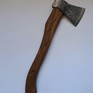 Peut inclure: Hache avec un manche en bois et une tête argentée. La tête présente un motif gravé avec des motifs détaillés et une figure centrale. Le manche en bois est incurvé et présente un fini veiné naturel.