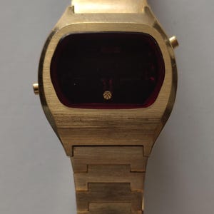 May include: A gold-tone digital wristwatch with a brushed metal finish, featuring a rectangular case and a matching link bracelet. The watch face has a dark red display screen with a small circular logo centered at the bottom of the screen.