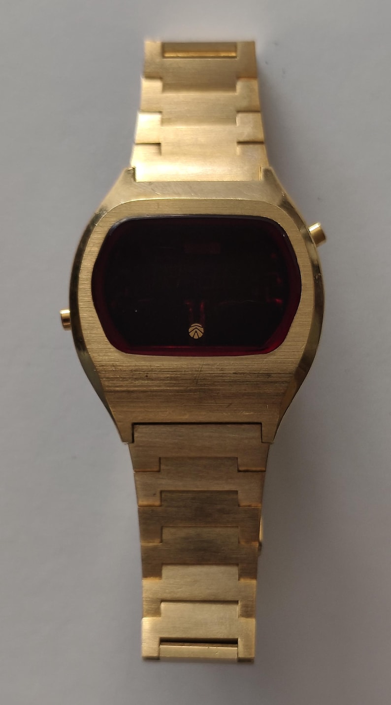 May include: A gold-tone digital wristwatch with a brushed metal finish, featuring a rectangular case and a matching link bracelet. The watch face has a dark red display screen with a small circular logo centered at the bottom of the screen.