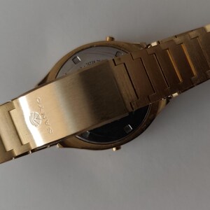 May include: A gold-toned wristwatch with a metallic link band. The clasp features the SANYO brand name and logo engraved on the surface. The watch is shown from the back, revealing a circular metal case back with visible text and small buttons.