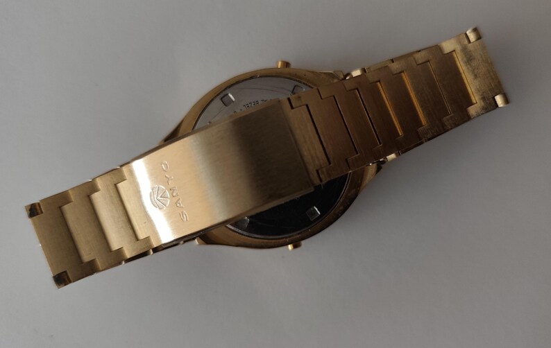 May include: A gold-toned wristwatch with a metallic link band. The clasp features the SANYO brand name and logo engraved on the surface. The watch is shown from the back, revealing a circular metal case back with visible text and small buttons.