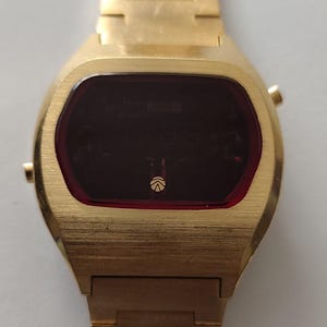 May include: A gold-tone digital wristwatch featuring a brushed metal finish, a rectangular case with rounded edges, and a dark red display screen. The watch face includes a small circular logo at the bottom center and two side buttons on the right.